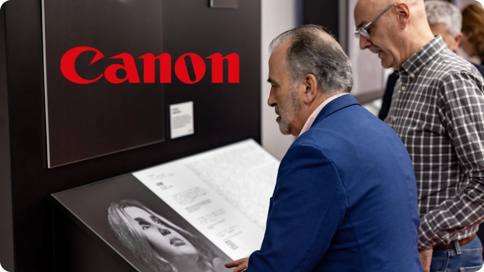 dos personas de mediana edad en una exposición, y uno de ellos con discapacidad visual que esta comprendiendo el contenido del descriptivo con braille y en el fondo aparece el logotipo de "Canon"